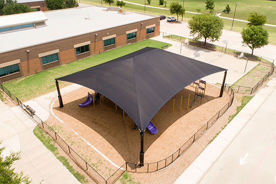 Ariel view of a large shade structure installation in the middle of a school playground. Underneath you can just see playground equipment, fully shaded.